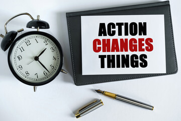 On the table there is a clock, a pen, a notebook and a card on which the text is written ACTION CHANGES THINGS