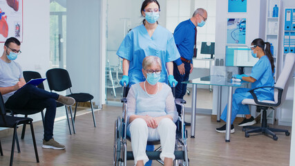 Senior man discussing with nurse at hospital reception wearing mask against coronavirus, assistant with sterile gloves is pushing disabled mature woman in wheelchair through waiting area.