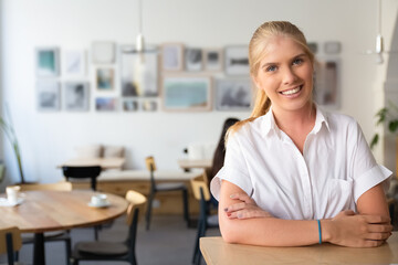 Happy beautiful blonde woman wearing white shirt, standing in co-working space, leaning on desk,...