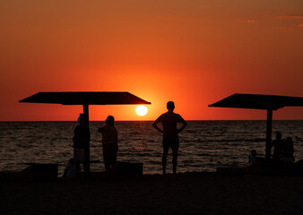Black silhouettes of people and beach umbrellas against a bright sunset.