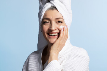Happy mature woman with head wrapped in towel applying anti-aging cream on clean skin