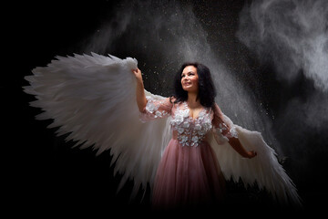 Girl with white wings posing in dark black studo during photoshoot with flour or dust and light. White angel in dark space