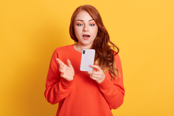 Stunned European woman being in shock, reads terrible news from cellular gadget, has wavy red hair, wears orange sweater, poses against yellow wall.
