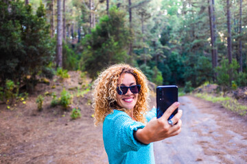 Cheerful beautiful caucasian woman adult take selfie picture in the forest enjoying trees and nature outdoor lesiure activity around - concept of tourism people