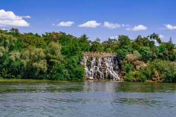 Fototapeta na wymiar Roaring Threshold Waterfall on the slope of the Monastery Island in Dnipro (Ukraine). Artificial decorative waterfall on the Dnieper river against the background of a green summer park