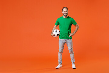 Fototapeta na wymiar Full length portrait smiling young man football fan in basic green t-shirt cheer up support favorite team with soccer ball isolated on orange background studio. People sport leisure lifestyle concept.