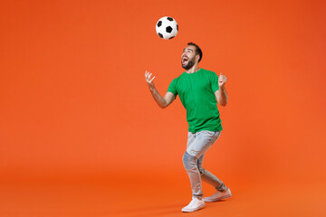Fototapeta na wymiar Full length portrait excited young man football fan in green t-shirt cheer up support favorite team throwing soccer ball isolated on orange background studio. People sport leisure lifestyle concept.