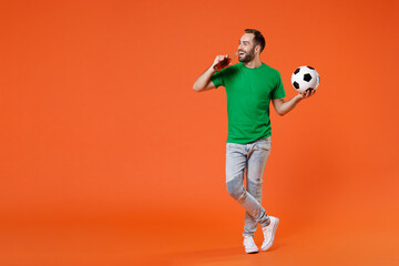 Fototapeta na wymiar Full length portrait cheerful young man football fan in green t-shirt cheer up support favorite team with soccer ball hold beer bottle isolated on orange background. People sport leisure concept.