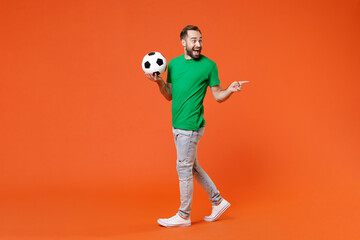 Fototapeta na wymiar Full length portrait excited man football fan in green t-shirt cheer up support favorite team with soccer ball pointing index finger aside isolated on orange background. People sport leisure concept.