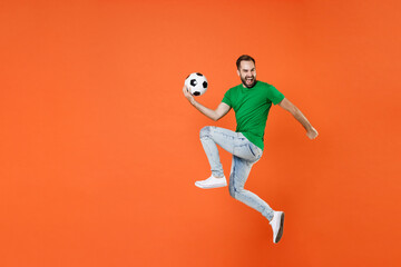 Fototapeta na wymiar Full length portrait of cheerful man football fan in green t-shirt cheer up support favorite team with soccer ball jumping like running isolated on orange background. People sport leisure concept.