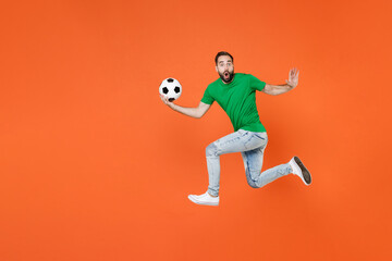 Fototapeta na wymiar Full length portrait of shocked man football fan in green t-shirt cheer up support favorite team with soccer ball jumping spreading hands isolated on orange background. People sport leisure concept.