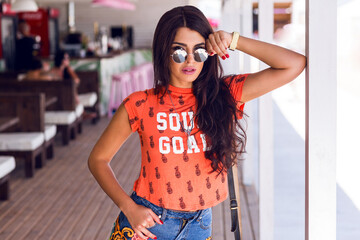 Outdoor summer fashion portrait of young long hair brunette posing in beach bar. She is smiling, enjoying  summer, wearing sunglasses, bright orange t-shirt and stylish denim  shorts. .