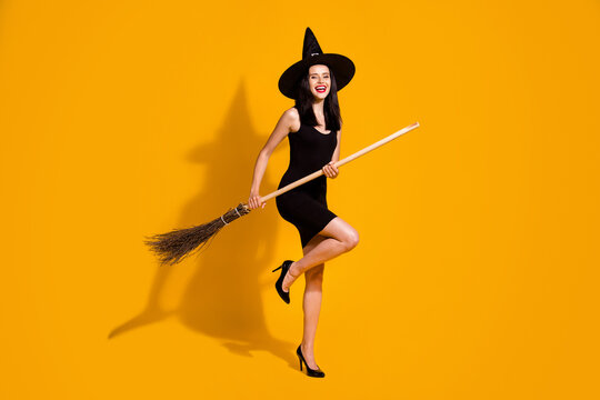 Full body size photo of pretty charming young sorceress lady smiling hold broomstick pretend sky flight wear black headwear high heels dress isolated bright yellow color background