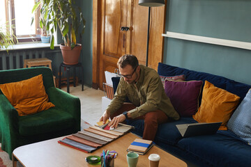 Fototapeta premium Mature designer in eyeglasses sitting on sofa in front of the table and working with fabric samples at modern office