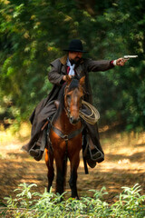The cowboy is on the horse, standing in the forest and holding a gun.