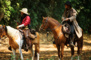 Two cowboy is on the horse, standing in the forest and holding a gun.