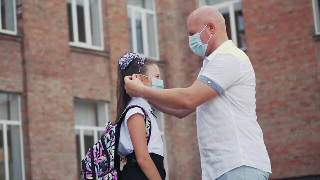 Dad puts protective, medical face mask on his daughter schoolgirl and gives small antiseptic spray, on school building background. Back to school after covid19 outbreak.