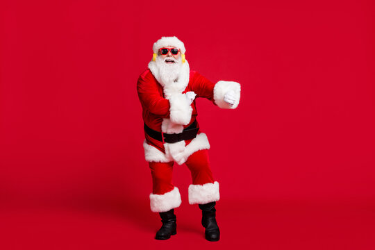 Full length photo of pensioner old man grey beard dance funny gesturing hand wear santa x-mas costume gloves coat belt sunglass headwear earphones black boots isolated red color background