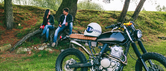 Young couple resting from a motorcycle trip having a beer outdoors with motorbike in foreground. Selective focus on couple in the background
