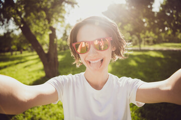 Closeup photo of carefree pretty lady make selfies sticking tongue out mouth social network blogger wear t-shirt sun specs spend free time having fun square garden green park outside