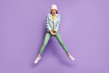 Full body photo of funky pretty lady stylish clothes jump high rejoicing good mood wear sun specs casual hat modern blue jacket green pants footwear isolated purple color background