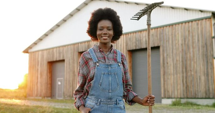 Portrait of beautiful African American young woman with pitchfork farmer standing in field at shed on background and smiling to camera. Pretty female with smile and fork at farm. Countryside concept. - Powered by Adobe