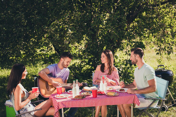 Photo of four best guys couple having gathering summer time fun sit table enjoy feast man play guitar serenade song friends listen hold drink punch beer cup under tree outside