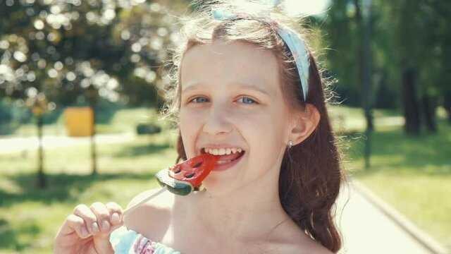 A satisfied girl with a bandage on her head is eating a candy on a stick shaped like a slice of watermelon. Rest on a day off in the park outside. Slow motion.