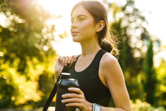 Image of young athletic sportswoman holding water bottle at summer park