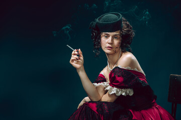 Smoking. Young woman as Anna Karenina isolated on dark blue background. Retro style, comparison of eras concept. Beautiful female model like literature character, great, old-fashioned.