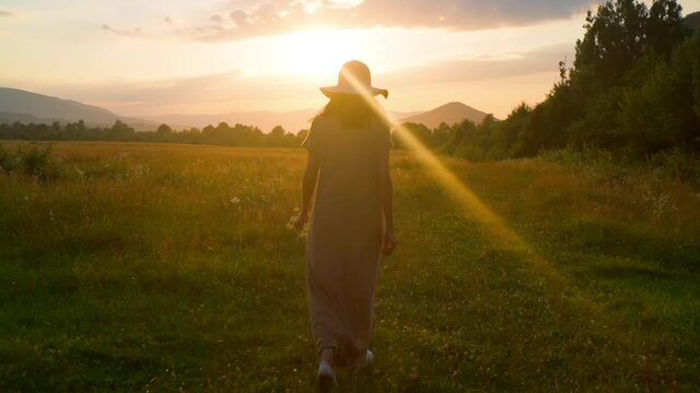 woman in maxi dress enjoy mountains landscape walks on the meadow with chamomiles happy summer vacation sky at sunset slow motion