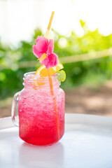 Cold red lemon soda drink with pink flower and lemon slice on glass jar at beach cafe. Fresh summer cocktail beverage concept