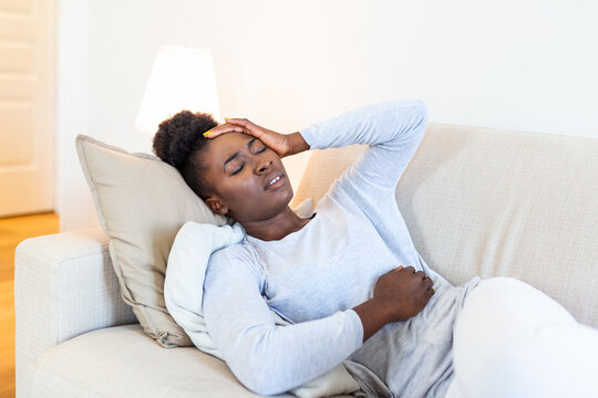 Portrait of a young black girl sitting on the couch at home with a headache and stomach pain. Beautiful woman suffering from chronic daily headaches. Sad woman holding her head because sinus pain