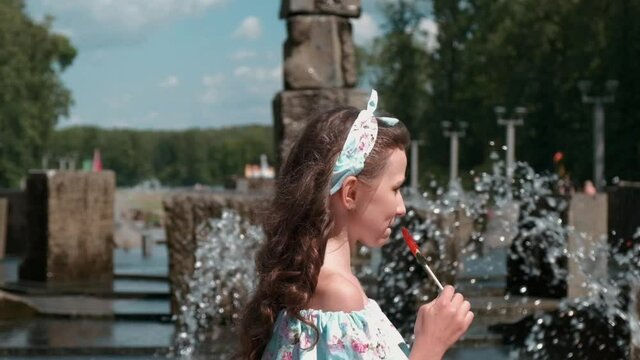 A beautiful girl in a full-length dress near the fountain. There is a sweet candy on a stick. Long beautiful hair. Rest on a day off in the park outside. Slow motion.