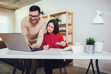 Happy couple doing business together working at small office on the laptop. They are using credit card for online shopping.