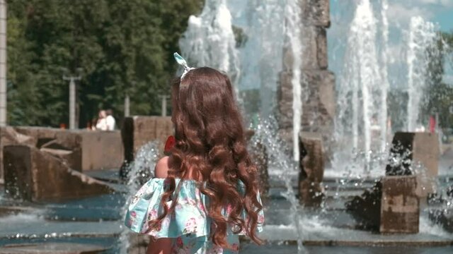 A beautiful girl in a full-length dress near the fountain. Candy on a stick in hand. Long beautiful hair. Rest on a day off in the park outside. Slow motion.
