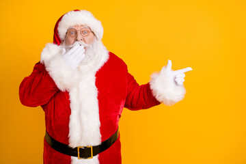 Photo astonished overweight santa claus impressed magic miracle x-mas adverts point index finger copyspace close cover lips hands wear headwear cap isolated bright shine color background