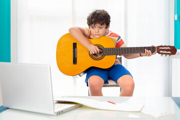 Little boy at home playing the Spanish guitar in front of his laptop. Online guitar class concept.