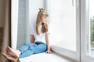 Little cute girl in a white T-shirt and jeans sits near the window
