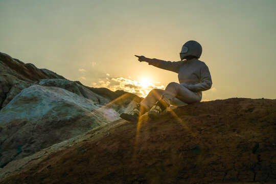 Side view of spaceman pointing at beautiful sky with stars while sitting on top of mountain. Mission specialist astronaut wearing white space suit with helmet. Concept of space travel and night sky - Powered by Adobe