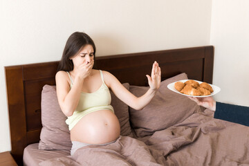 Obraz na płótnie Canvas Pregnant woman relaxing in bed makes stop gesture to croissants. Expecting mother refuses to eat pastry. Diet during pregnancy concept
