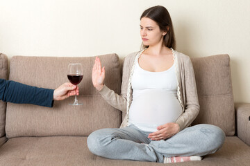 Pregnant woman sitting on the sofa refuses to drink wine and makes stop gesture to the glass. No alcohol during pregnancy concept