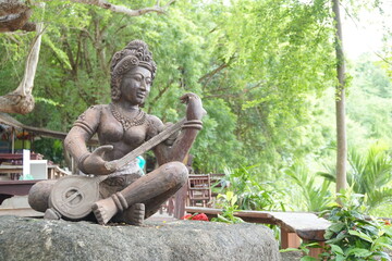 Statue of a woman playing the Thai harp on a rock and the background is a tree. It is a traditional musical instrument of Thailand.