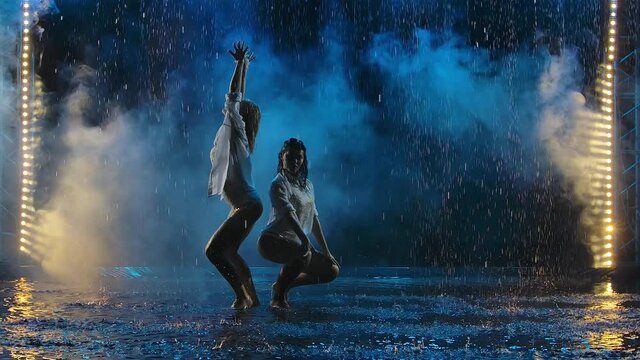 Two young women passionately dancing salsa elements in raindrops and smoke. Female silhouettes in wet bodysuits move erotically in slow motion.