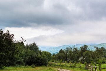 Nature in the village. Trees, mountains in the background in the fog. Green and blue colors. The fence and the road are brown. Calm and rest. Clouds. Gloomy sky.