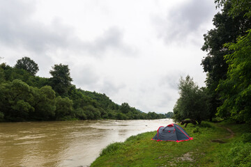 Tent by the river. Gloomy sky and fog in the distance. Dirty brown water. Trees in the valley. Green grass. Leisure and travel. Camping. Motivation.