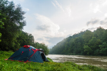 Tent by the river. Gloomy sky and fog in the distance. Dirty brown water. Trees in the valley. Green grass. Leisure and travel. Camping. Motivation.