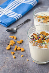 Top view of two glasses with natural yogurt, cookie pieces, colors, on gray background with spoon, cookies and blue napkin, in vertical