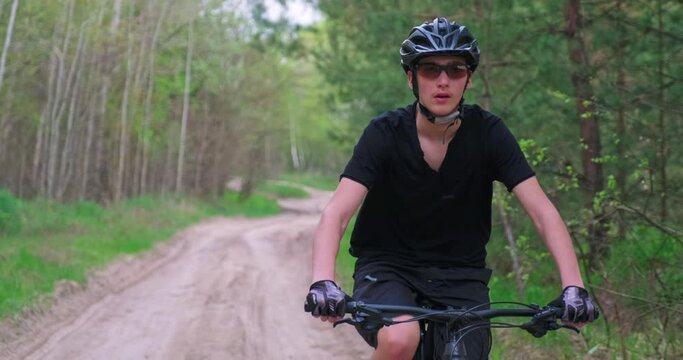 Front view to the man in a Bicycle helmet riding an mtb bike in the summer.Young  man cyclist rides on a forest road. Teenager on a mountain bike rides through the forest. Real time footage