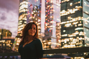 A young woman stands at night in front of glowing glass skyscrapers in Moscow. High ISO, grainy and noisy images.